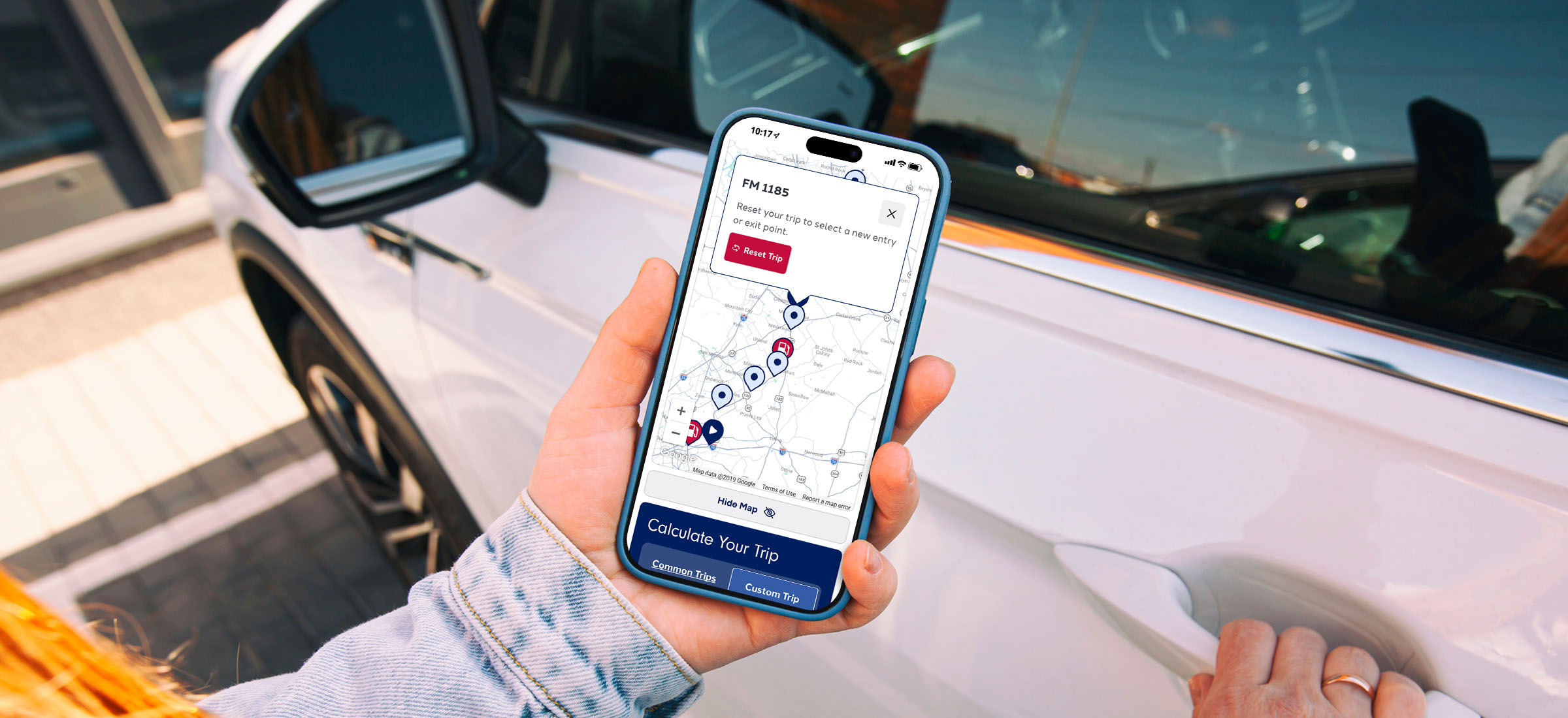 A hand holding a phone showing the SH 130 Trip Planner map in front of a car.
