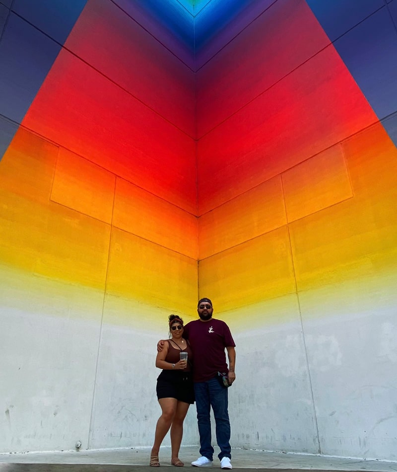 Alex standing in front of a rainbow wall with her partner.