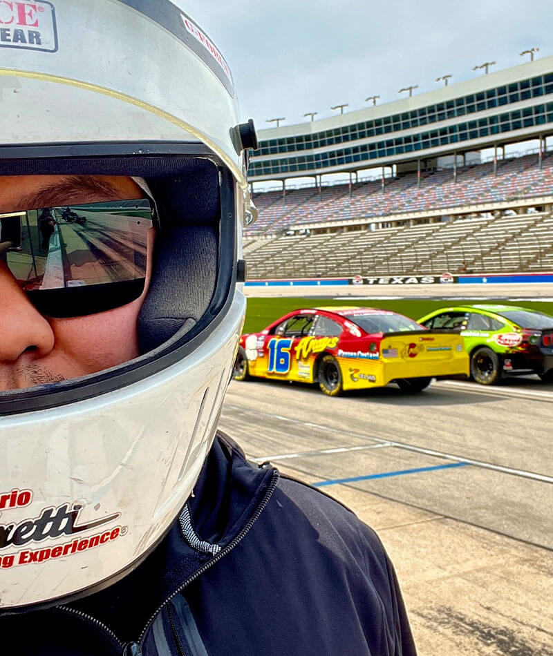 Patrick in a racing helmet stands on a racetrack with two colorful race cars behind, set against empty stands.