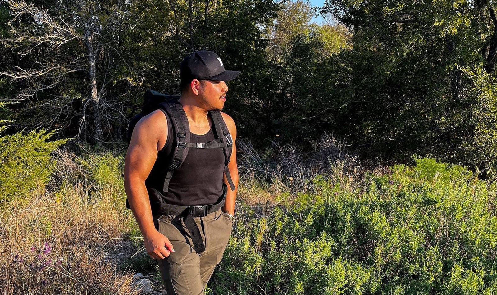 Patrick wearing a black tank top, cap, and backpack standing in a sunlit forest clearing, gazing to the side.